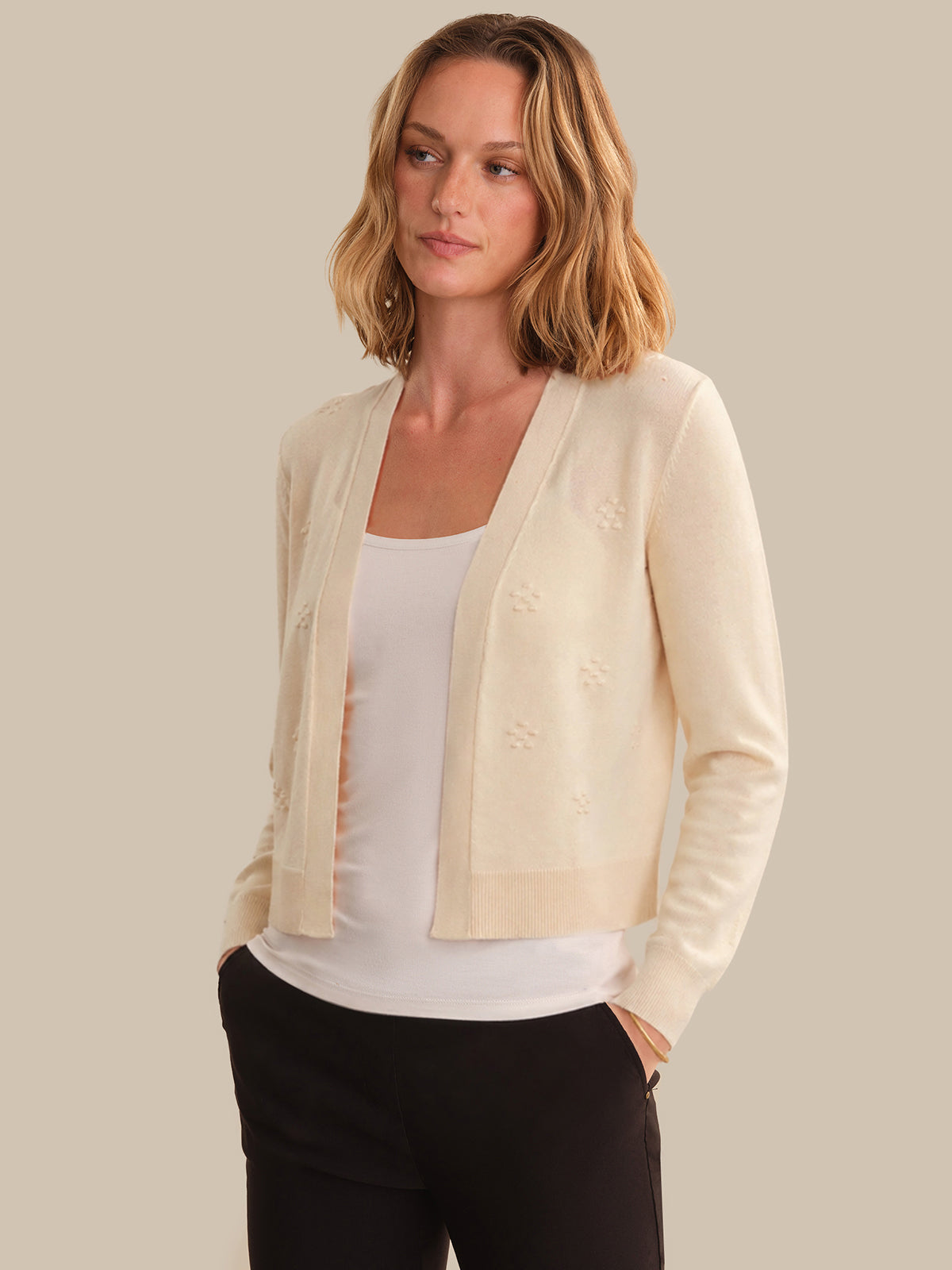 Woman wearing a light beige cardigan over a white top and black pants on a neutral background