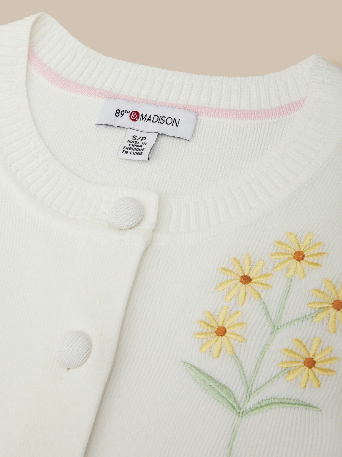 White cardigan with yellow floral embroidery and a visible brand tag on a beige background