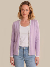 Woman wearing a light purple cardigan with lace trim over a white shirt and blue jeans on a beige background
