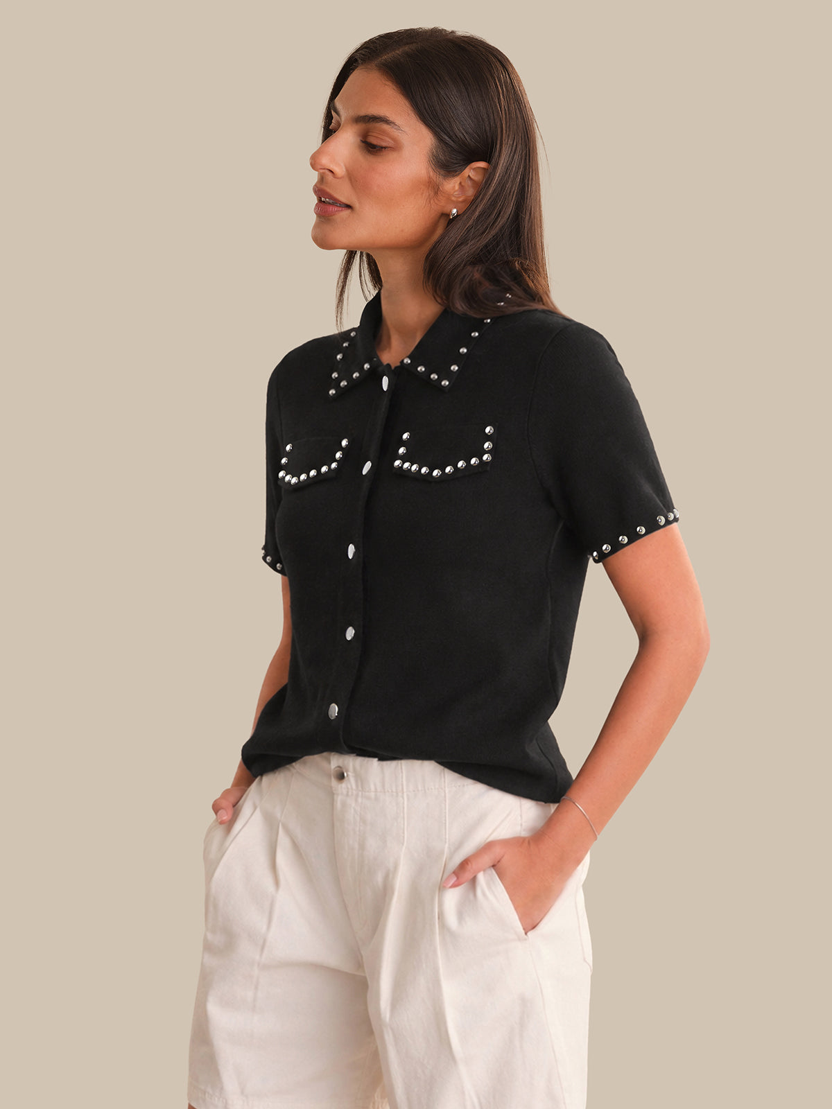 Woman wearing a black studded shirt with white pants on a beige background