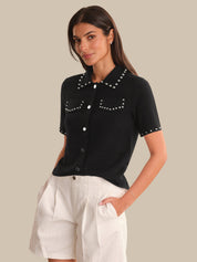 Woman wearing a black short-sleeve shirt with white buttons and trim, standing against a neutral background.