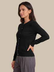 A woman modeling a black long-sleeve ribbed button-front cardigan with a round neckline, paired with gray trousers, against a solid beige background.