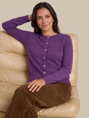 A woman wearing a purple long-sleeve button-front knit top and brown pants, seated on a beige leather couch against a plain beige background.