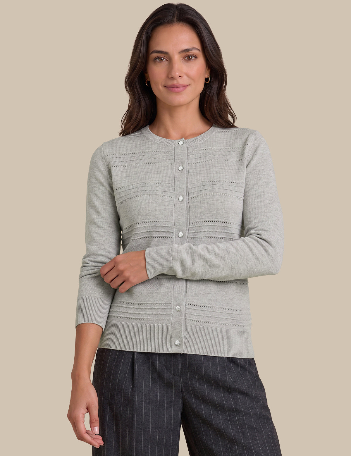 A woman modeling a light gray knit cardigan with button-front closure and textured horizontal ribbing, paired with dark pinstriped trousers, against a solid beige studio background.