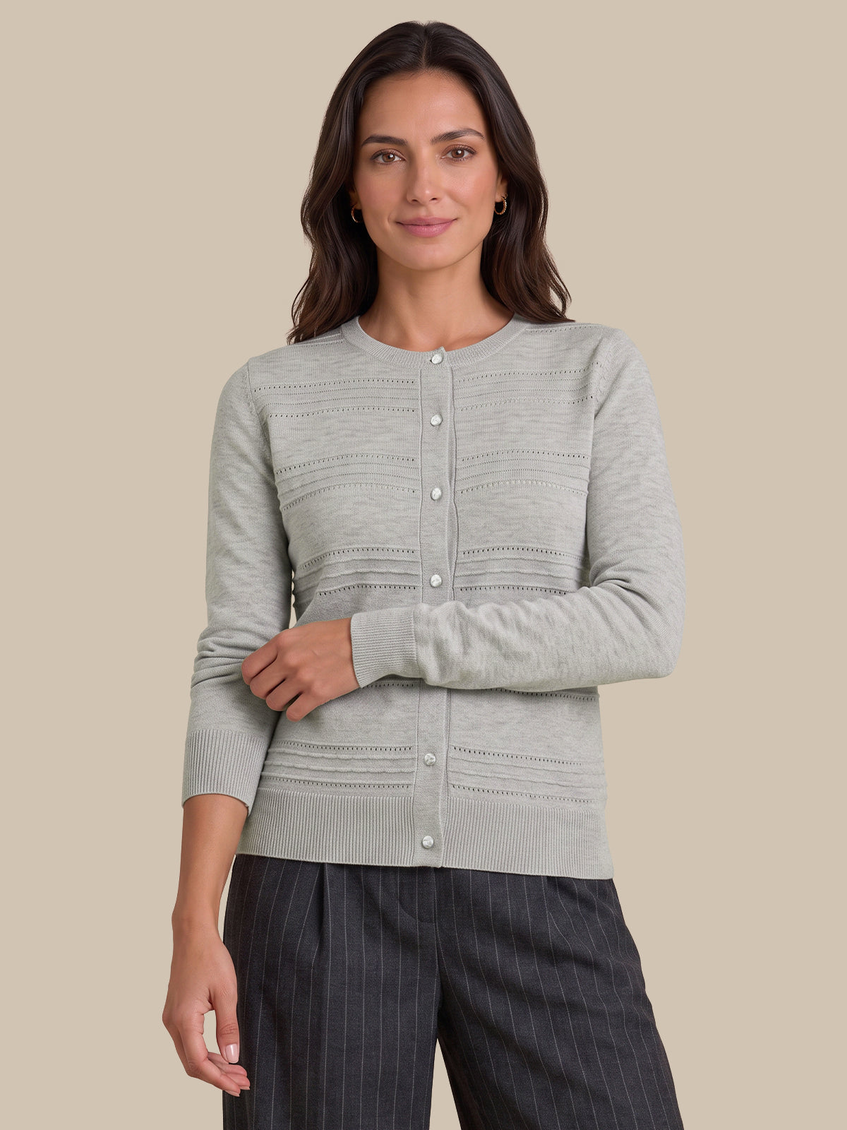 A woman modeling a light gray knit cardigan with button-front closure and textured horizontal ribbing, paired with dark pinstriped trousers, against a solid beige studio background.