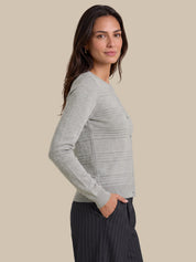 A woman wearing a light gray ribbed knit cardigan sweater and dark pinstriped trousers, shown in a side profile against a solid beige background.