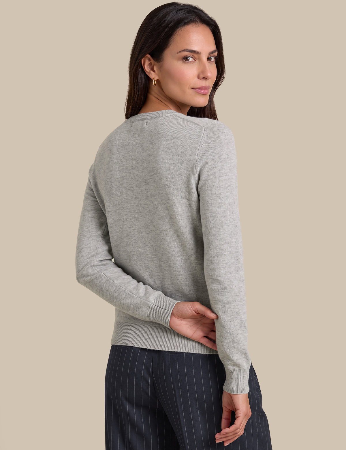 A woman shown from the back wearing a light gray knit sweater and dark pinstriped trousers, photographed against a plain beige studio background.