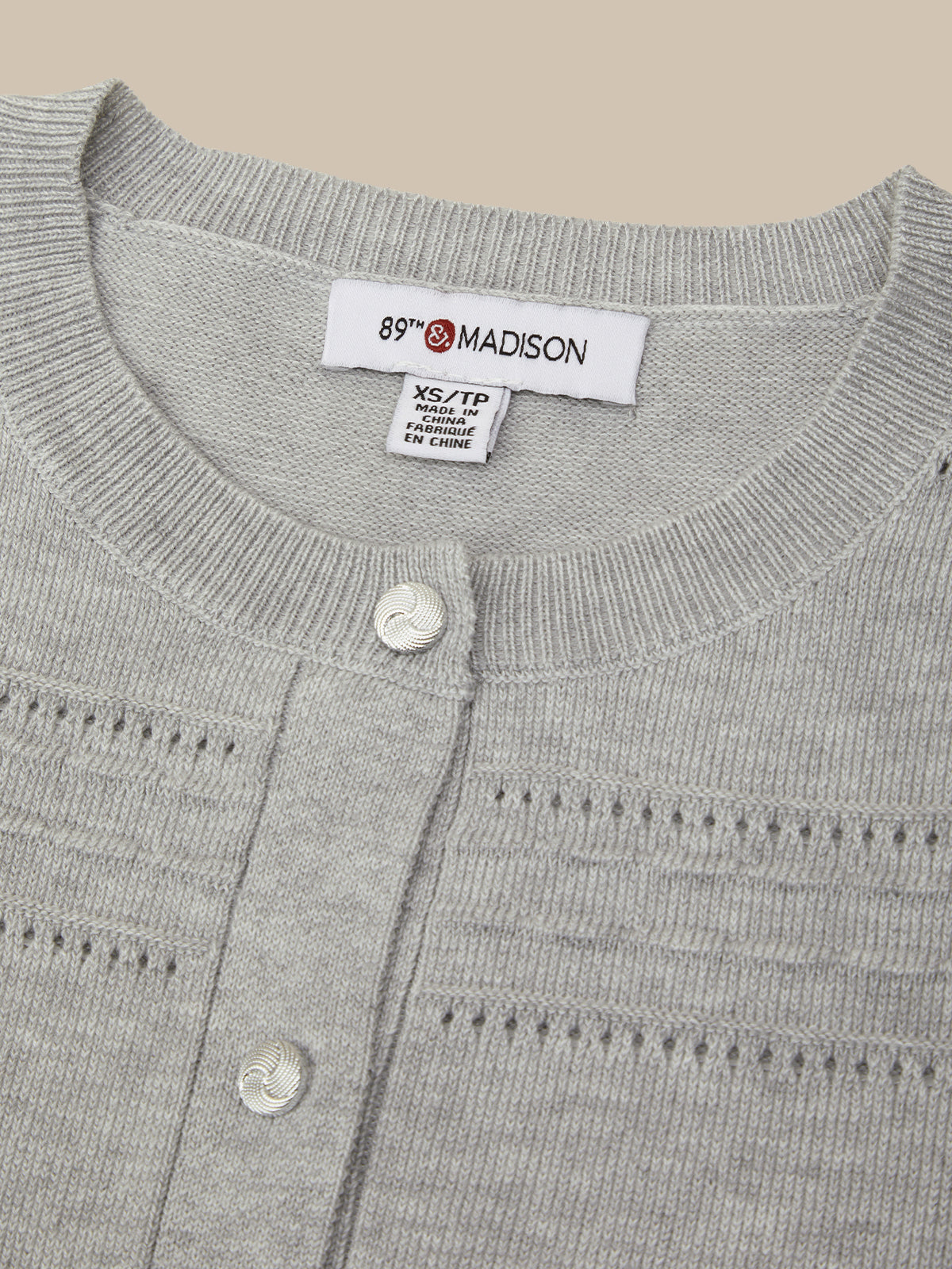 Close-up of a light gray knit cardigan sweater showing the inner neck label which reads '89th & MADISON' and 'XS/XXL', along with two white buttons on the front.
