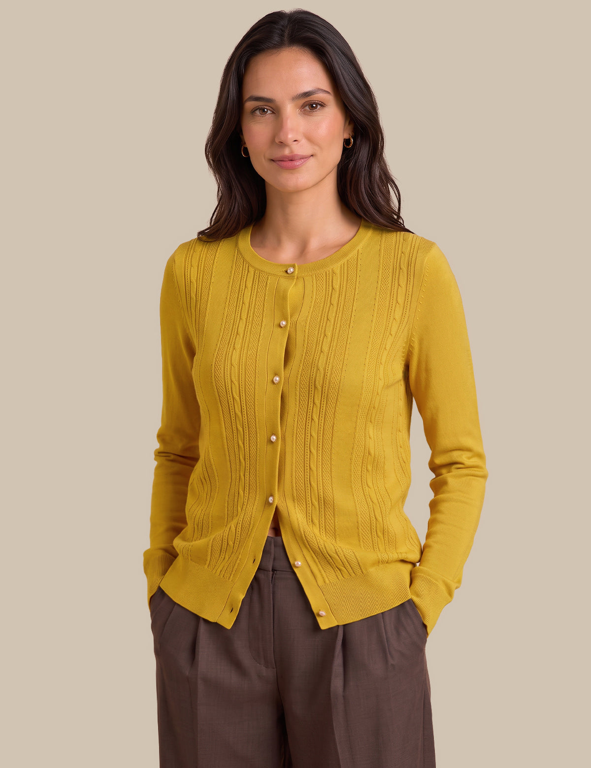 A woman modeling a mustard yellow ribbed button-front cardigan sweater with brown trousers, set against a solid beige studio background.
