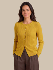 A woman modeling a mustard yellow ribbed button-front cardigan sweater with brown trousers, set against a solid beige studio background.