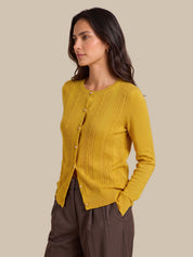 A woman wearing a mustard yellow ribbed knit cardigan with white buttons and brown trousers, photographed against a solid beige studio background.