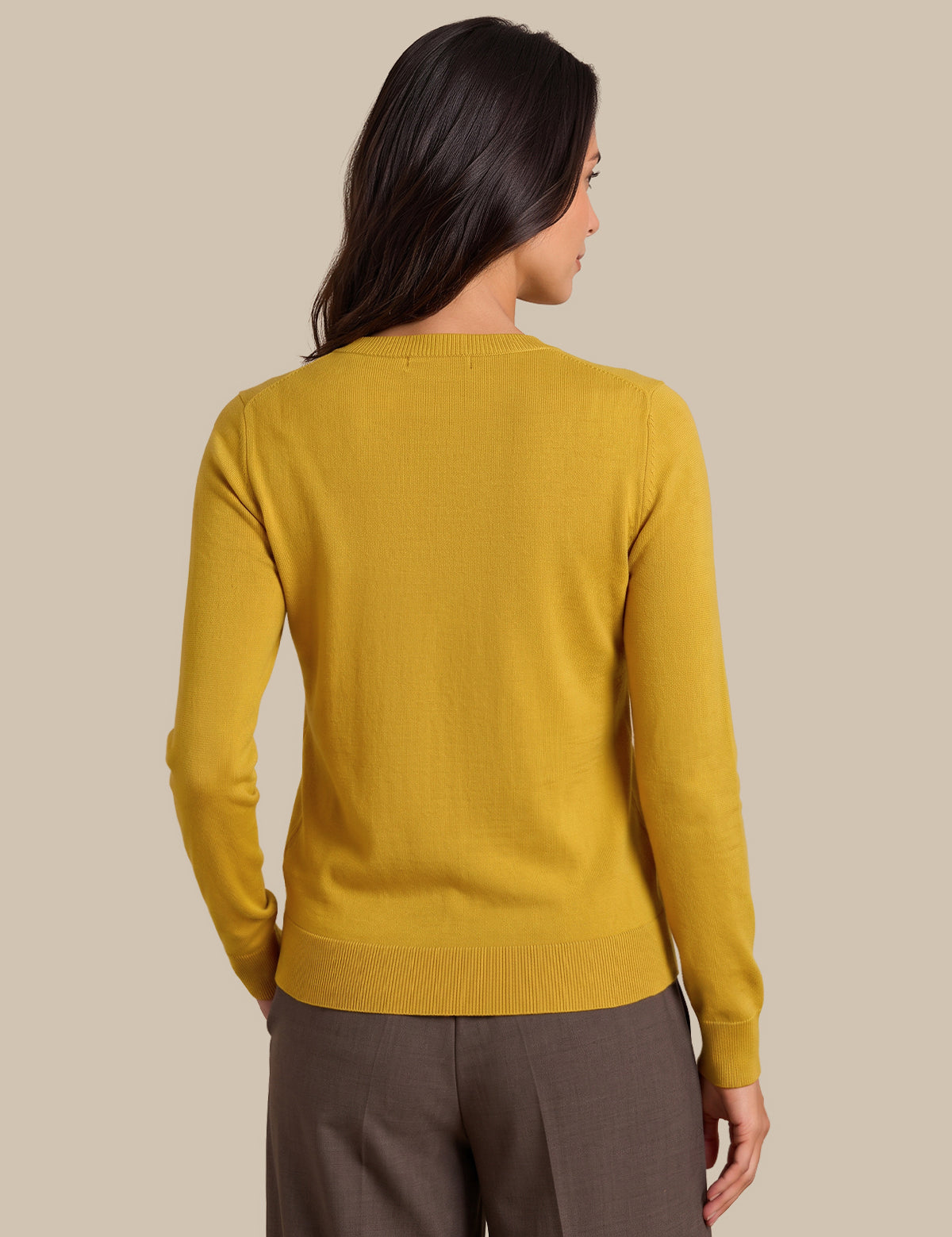 A woman shown from the back wearing a mustard yellow knit sweater and brown trousers, set against a solid beige studio background.