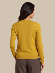 A woman shown from the back wearing a mustard yellow knit sweater and brown trousers, set against a solid beige studio background.
