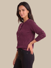 A woman modeling a long-sleeve ribbed knit cardigan in deep plum purple with white buttons, paired with black pants, against a solid beige background.