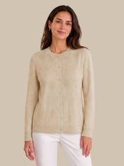 A woman wearing a light beige ribbed knit cardigan sweater with a button-up front, paired with white pants, against a solid beige background.