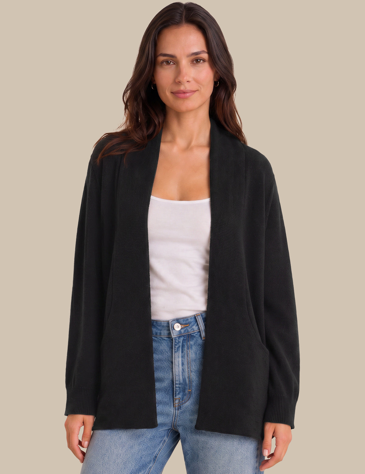 A woman modeling a black open-front cardigan over a white tank top and blue jeans, set against a solid beige studio background.