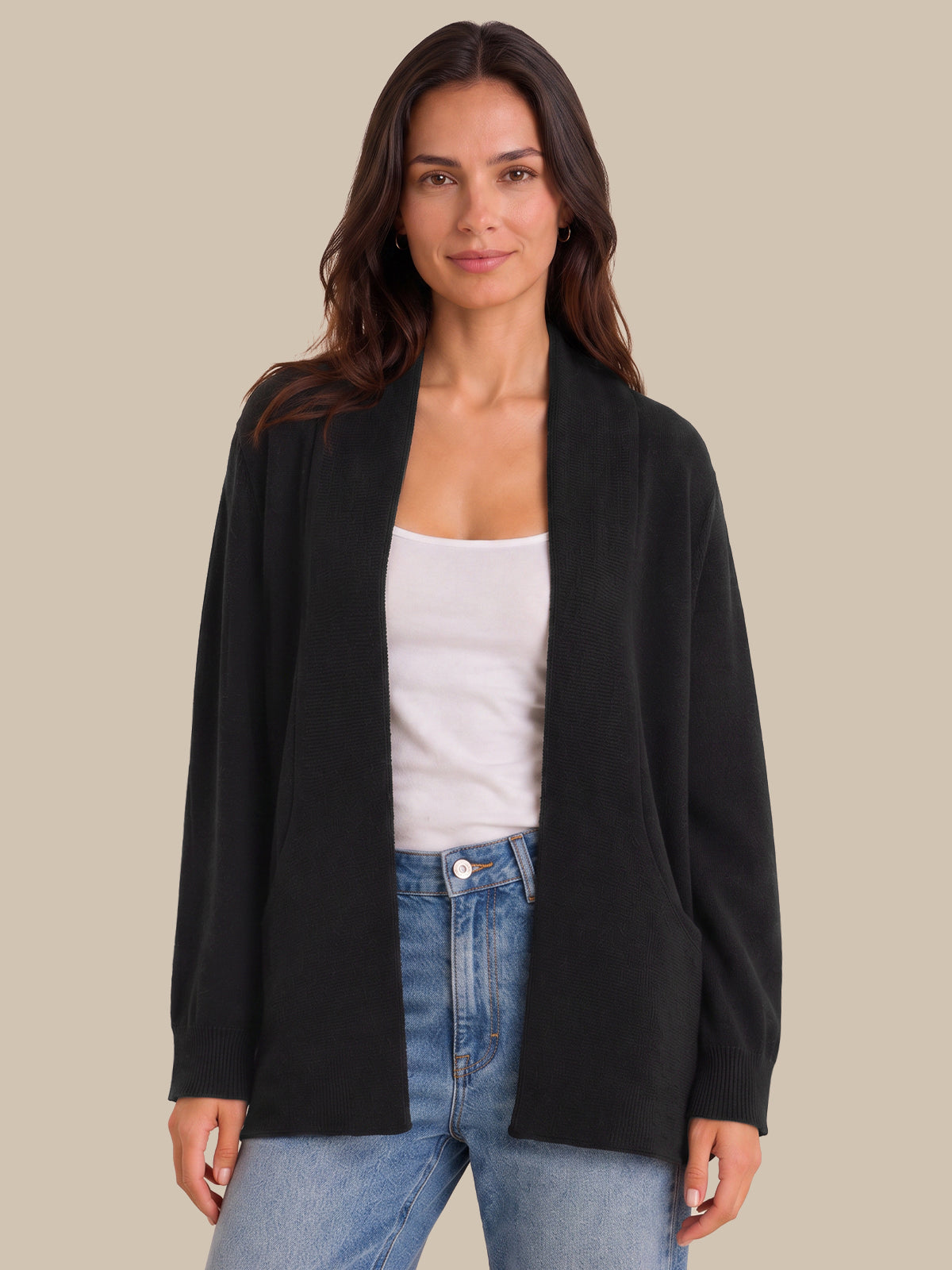 A woman modeling a black open-front cardigan over a white tank top and blue jeans, set against a solid beige studio background.