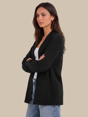 A woman modeling a black open-front cardigan over a white top and light blue jeans, standing against a solid beige background.
