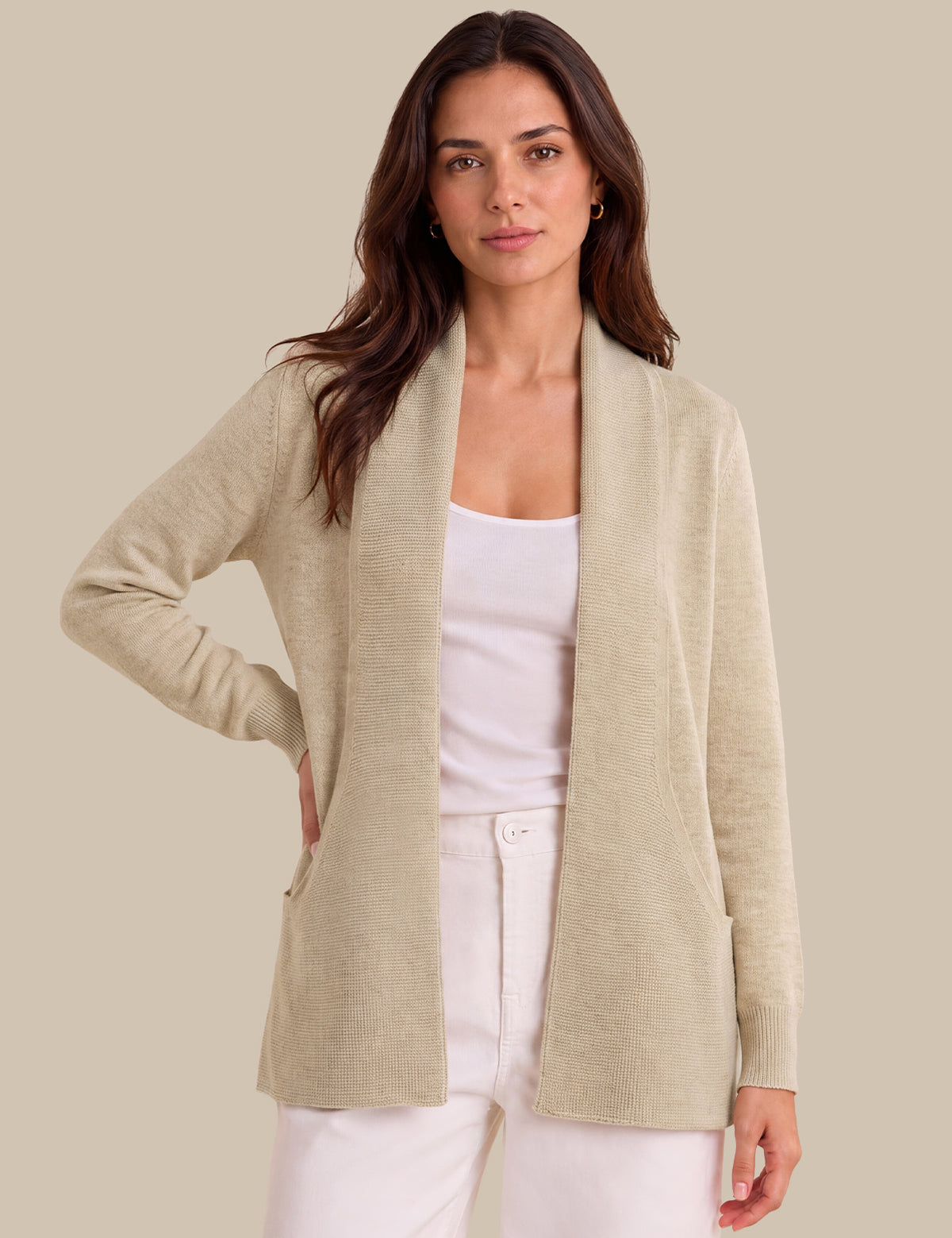 A woman modeling a light beige open-front knit cardigan over a white tank top and white pants, photographed against a plain warm beige studio background.
