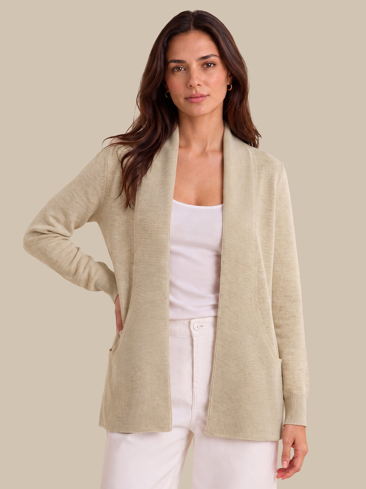 A woman modeling a light beige open-front knit cardigan over a white tank top and white pants, photographed against a plain warm beige studio background.