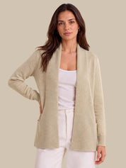 A woman modeling a light beige open-front knit cardigan over a white tank top and white pants, photographed against a plain warm beige studio background.