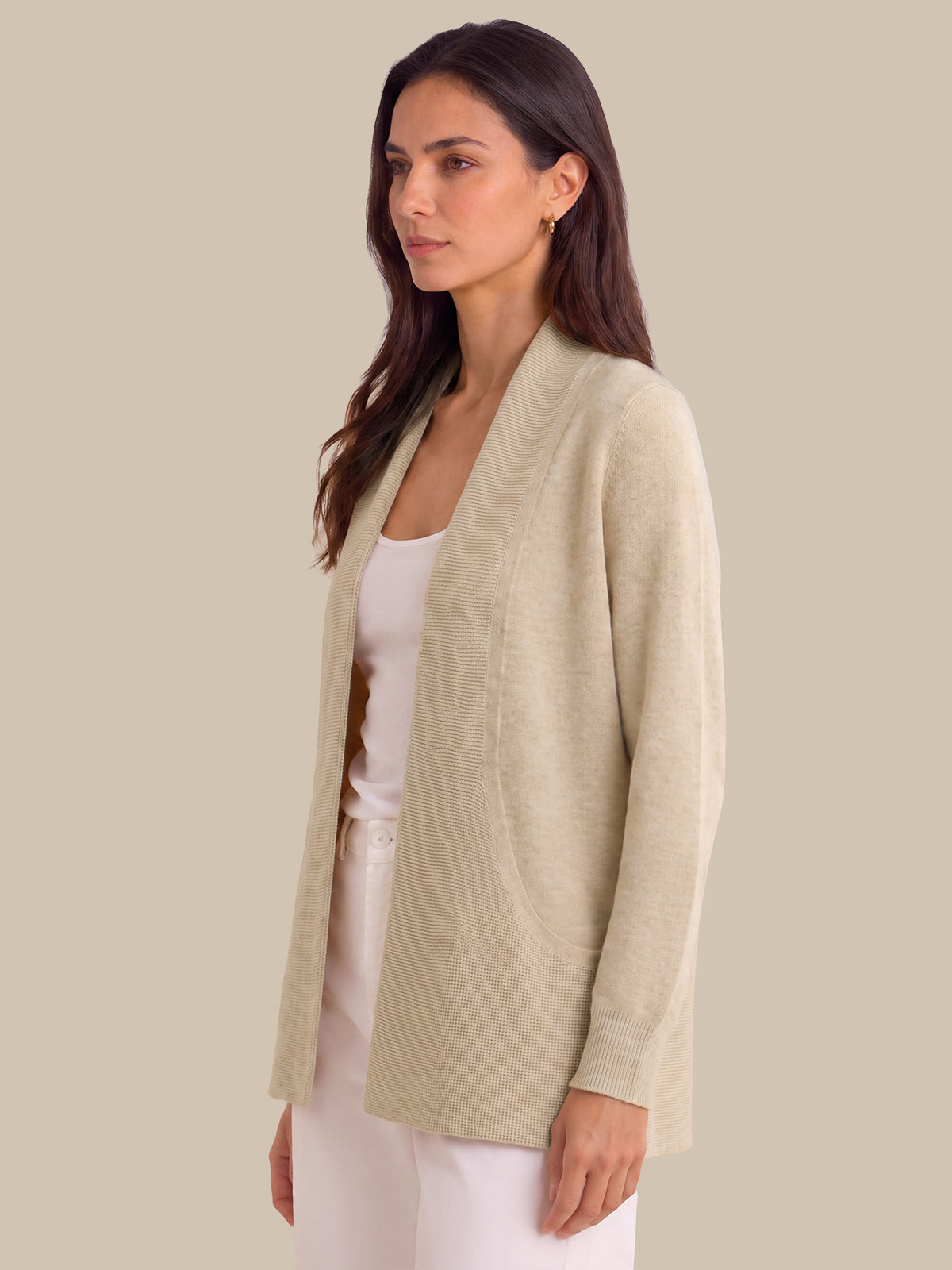 A woman wearing a light beige open-front cardigan over a white tank top and white pants, photographed against a solid beige studio background.