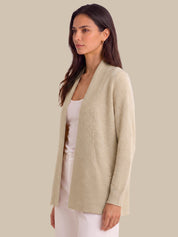 A woman wearing a light beige open-front cardigan over a white tank top and white pants, photographed against a solid beige studio background.