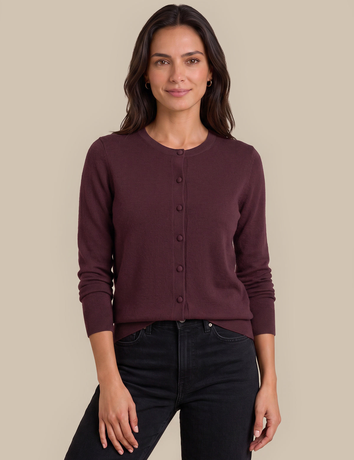 A woman modeling a dark burgundy button-up cardigan sweater with black jeans, standing against a solid beige studio background.