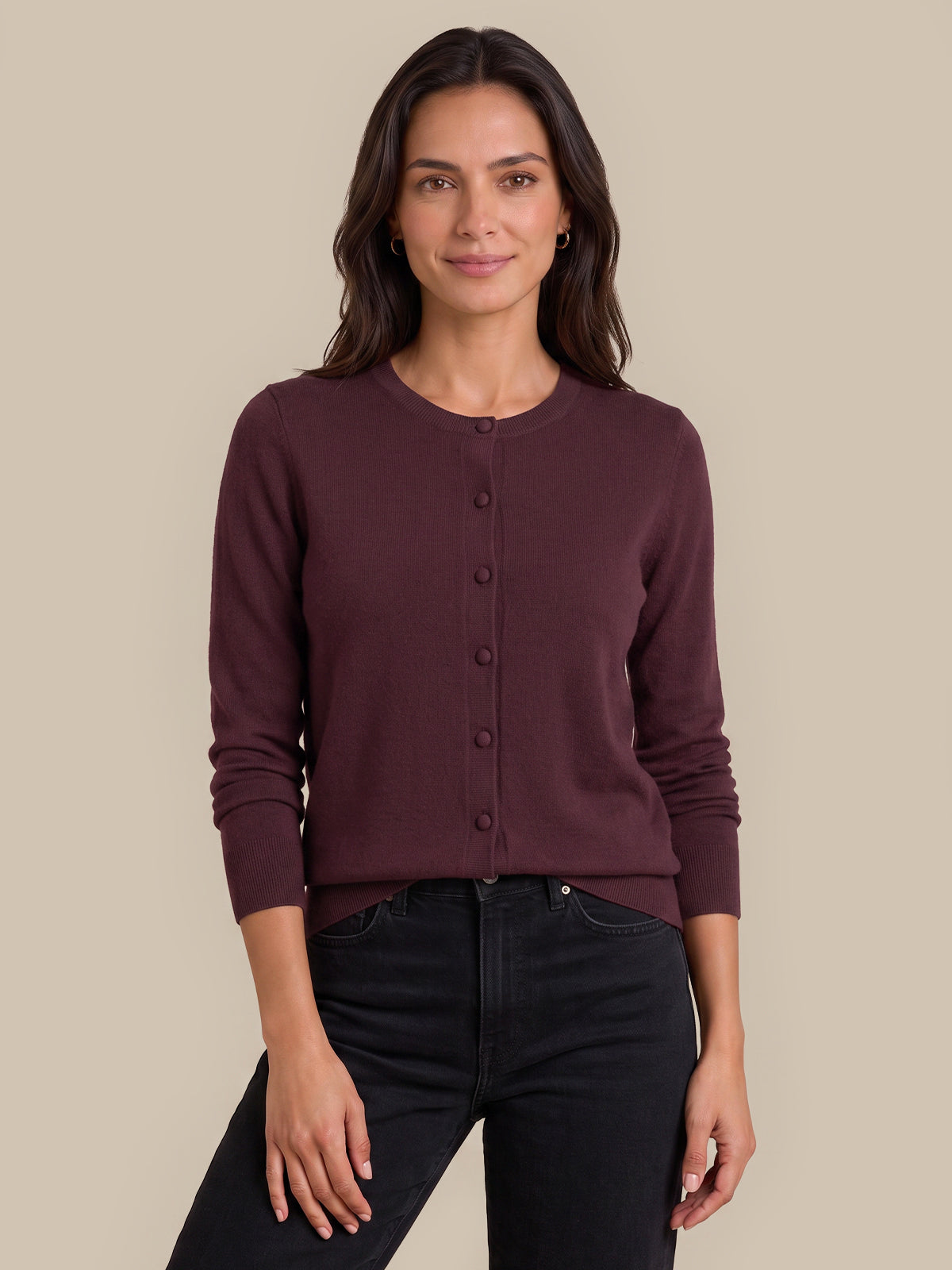 A woman modeling a dark burgundy button-up cardigan sweater with black jeans, standing against a solid beige studio background.