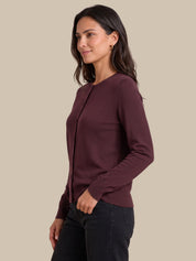 A woman wearing a long-sleeve crewneck knit top in deep burgundy, paired with dark jeans, shown in a side-profile view against a solid beige background.