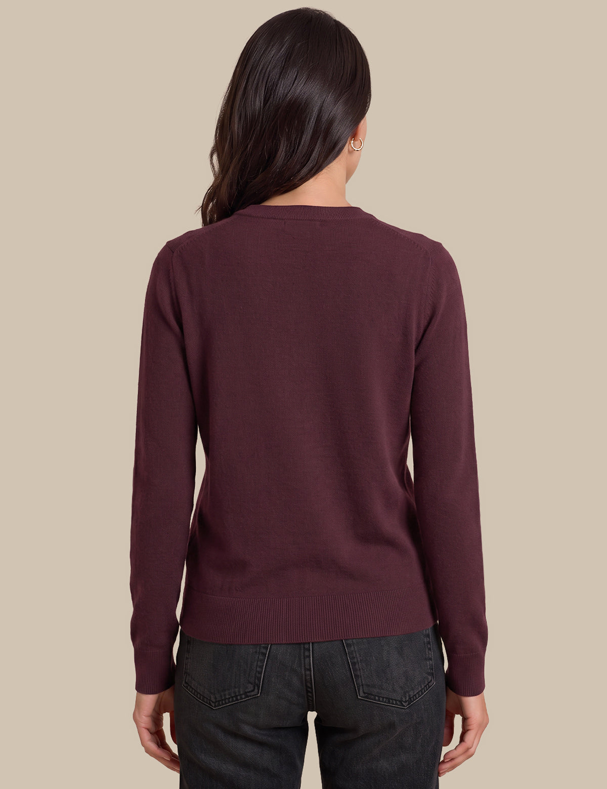 A woman viewed from the back wearing a long-sleeve crewneck sweater in a deep burgundy color, paired with dark gray jeans, against a solid beige background.