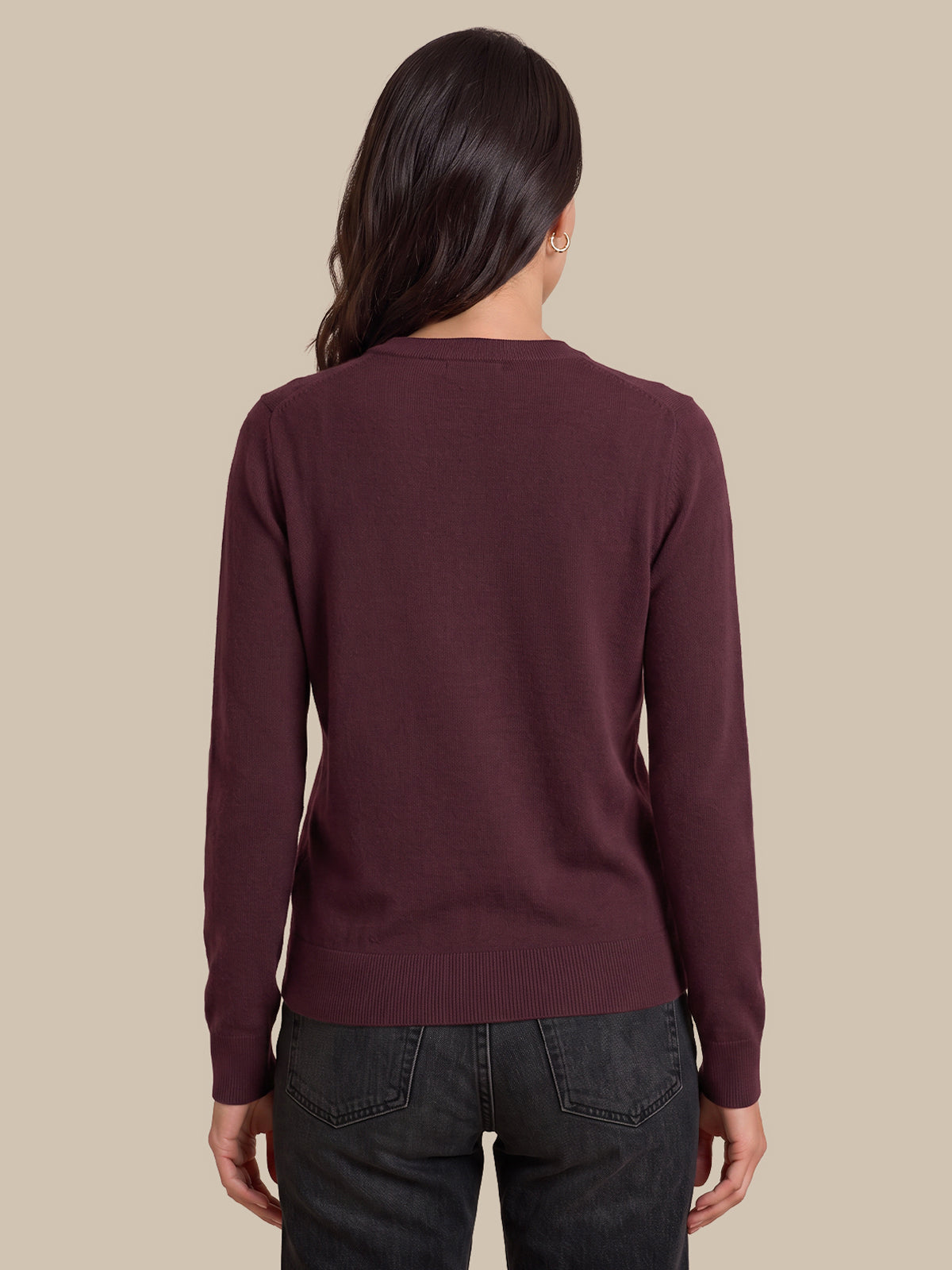 A woman viewed from the back wearing a long-sleeve crewneck sweater in a deep burgundy color, paired with dark gray jeans, against a solid beige background.