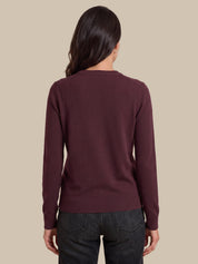 A woman viewed from the back wearing a long-sleeve crewneck sweater in a deep burgundy color, paired with dark gray jeans, against a solid beige background.