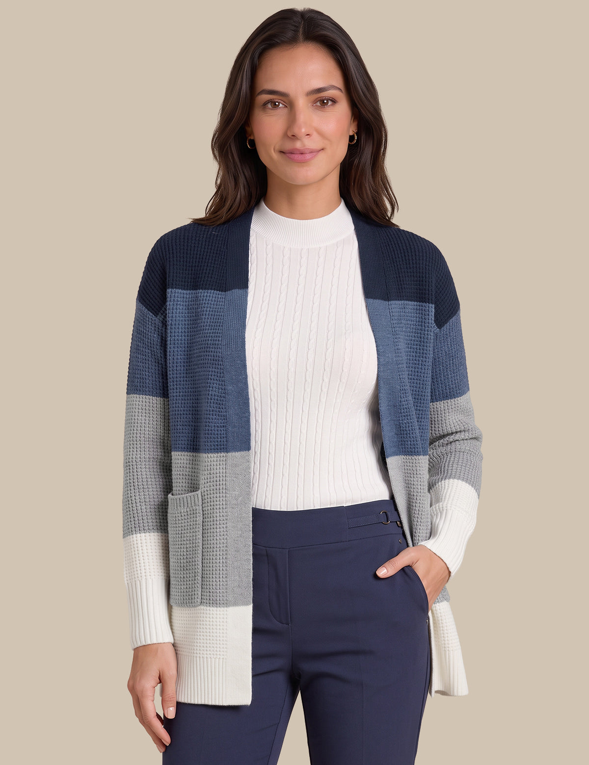 A woman modeling a color-block knit cardigan with navy, slate blue, gray, and cream sections, worn over a white ribbed top and navy pants against a solid beige background.