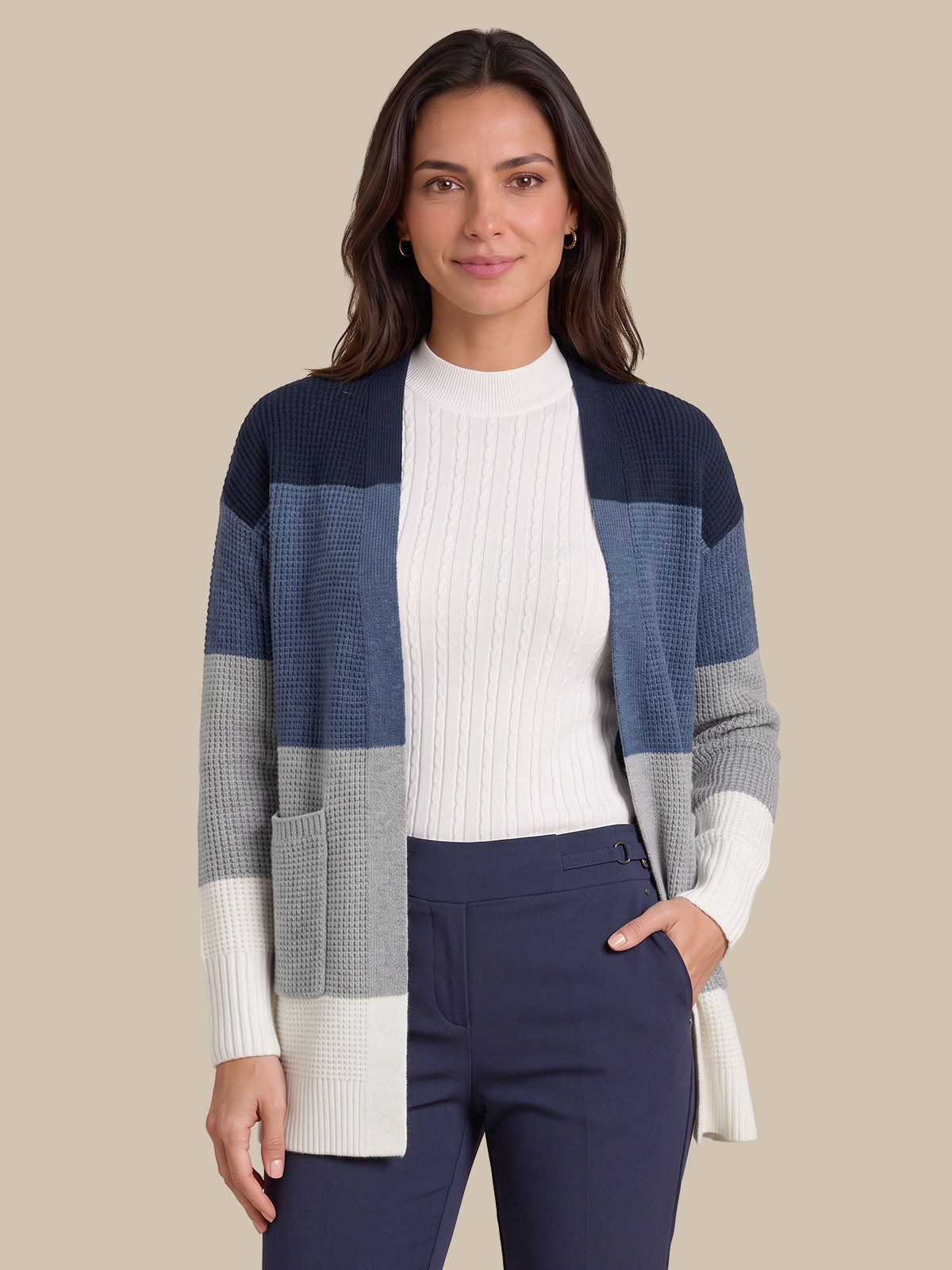 A woman modeling a color-block knit cardigan with navy, slate blue, gray, and cream sections, worn over a white ribbed top and navy pants against a solid beige background.