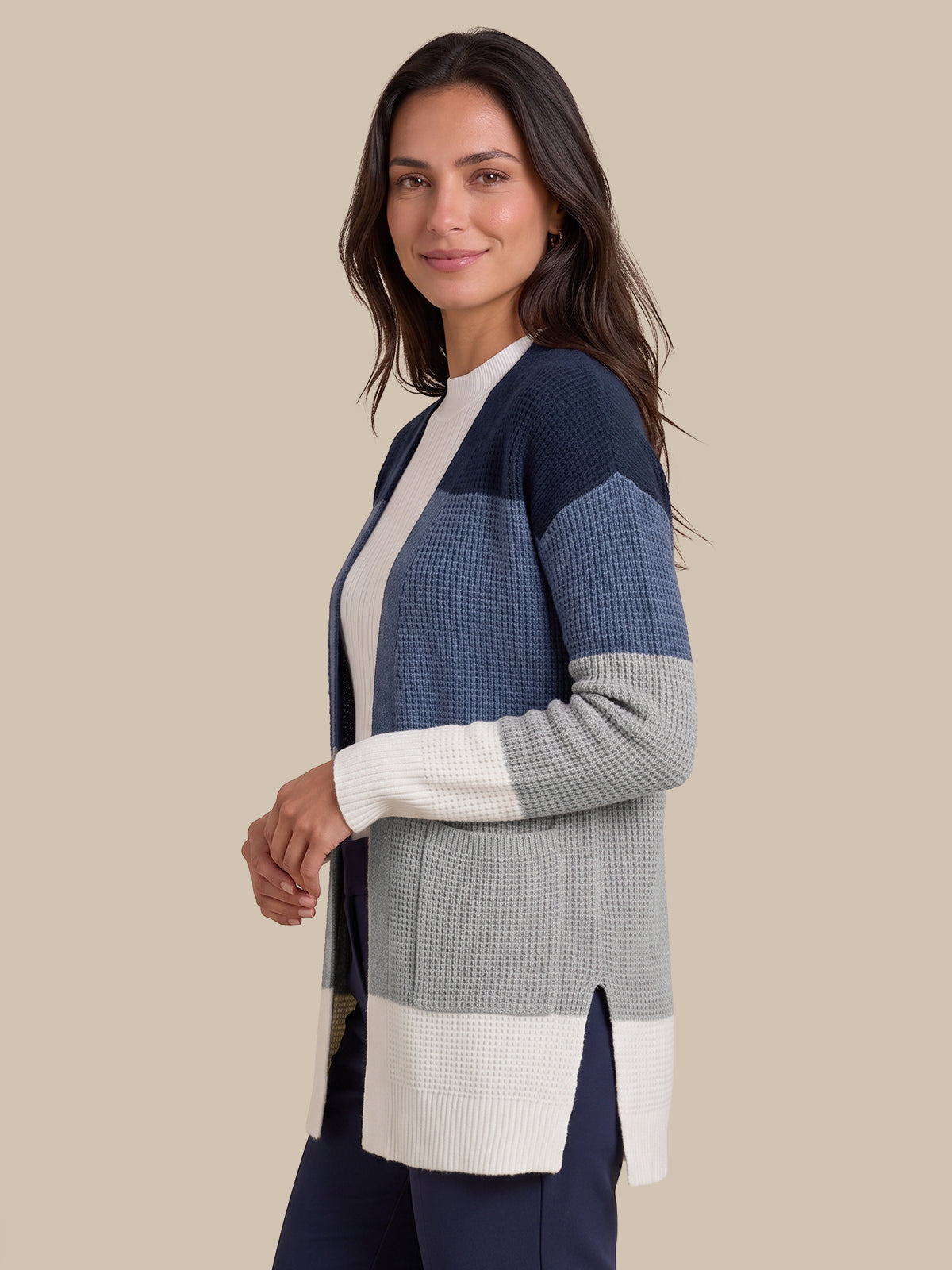 A woman modeling a color-block knit cardigan with navy blue, textured medium blue, light gray, and off-white sections, set against a solid beige background.
