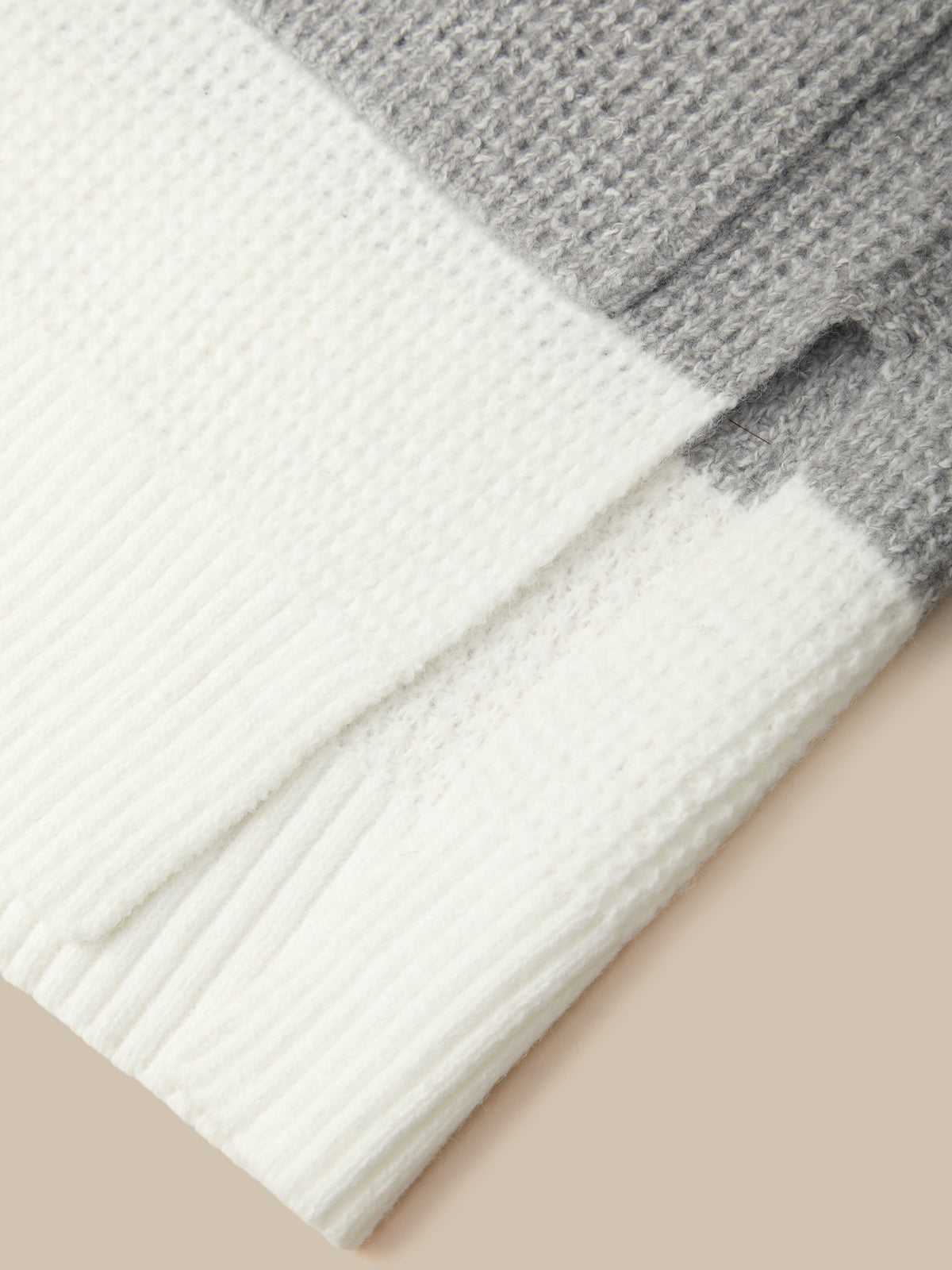 Close-up of a folded knit blanket or throw in white and gray waffle and ribbed textures on a beige background.