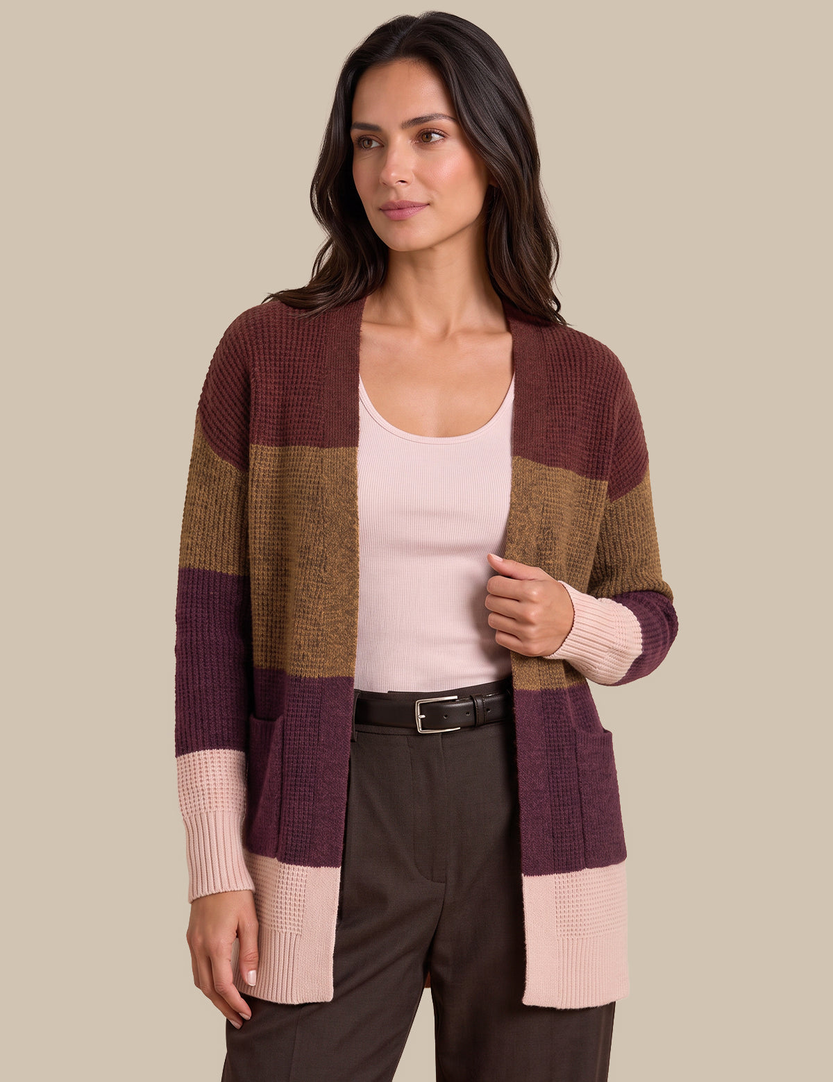 A woman modeling a color-block knit cardigan with horizontal stripes of burgundy, tan, and light pink, worn over a pale pink tank top and dark brown trousers against a solid beige background.