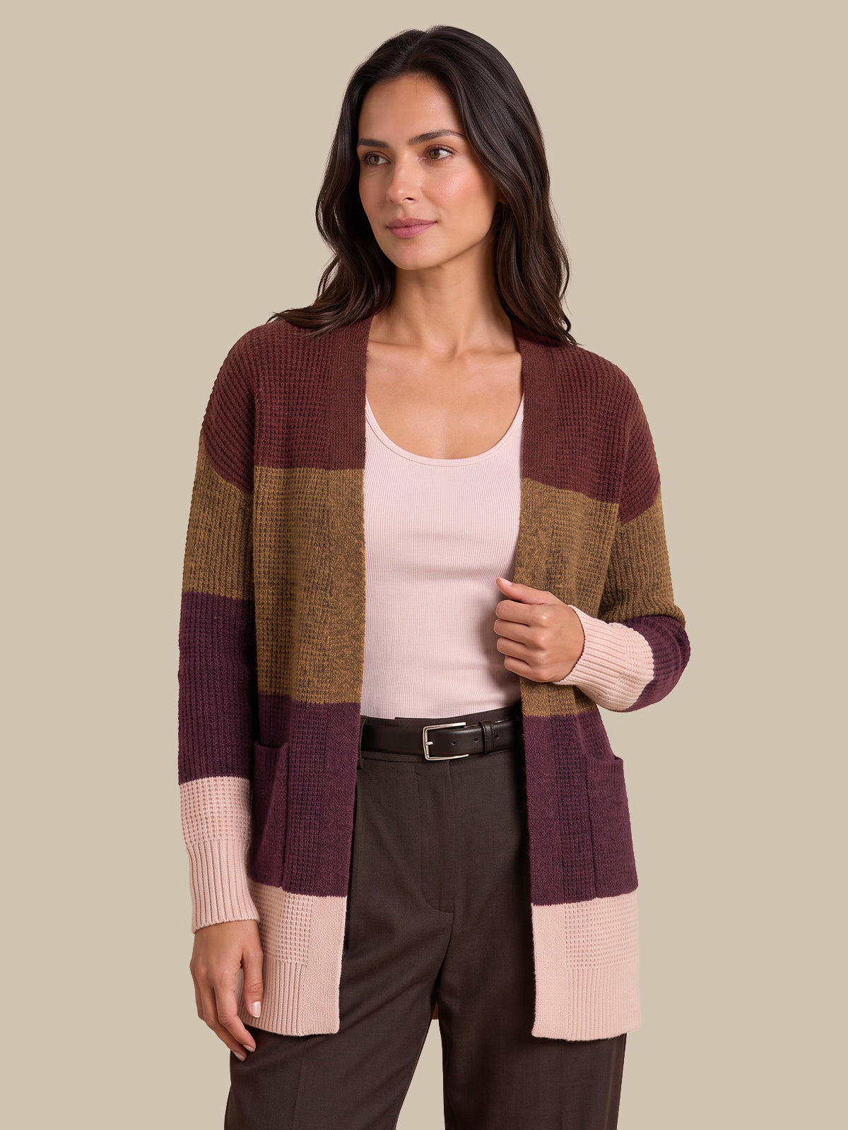 A woman modeling a color-block knit cardigan with horizontal stripes of burgundy, tan, and light pink, worn over a pale pink tank top and dark brown trousers against a solid beige background.