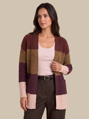 A woman modeling a color-block knit cardigan with horizontal stripes of burgundy, tan, and light pink, worn over a pale pink tank top and dark brown trousers against a solid beige background.