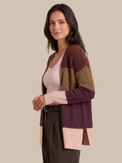 A woman modeling a color-blocked ribbed knit cardigan with sections of deep burgundy, olive green, and light pink, worn over a pale pink top and dark brown trousers against a solid beige background.
