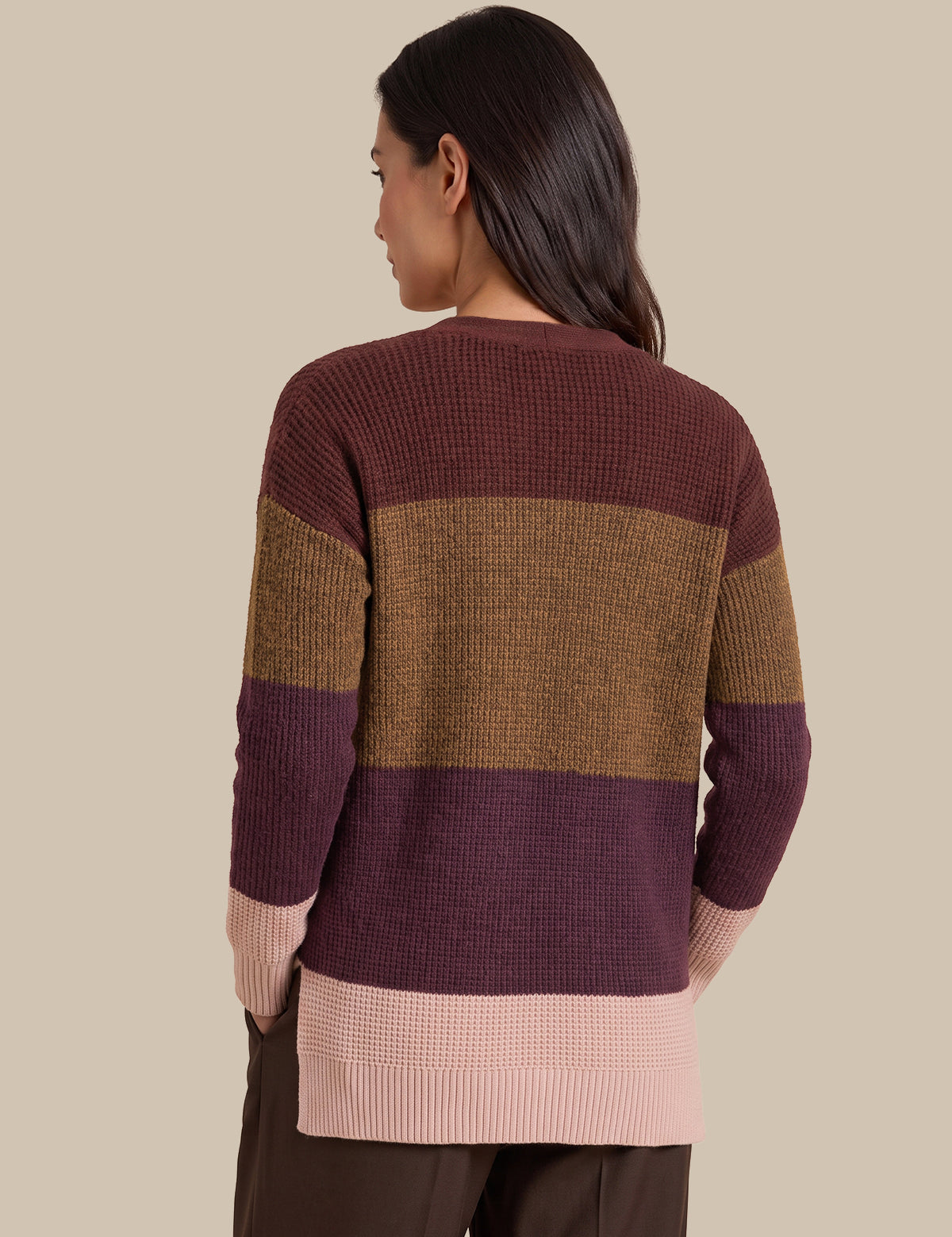 A woman shown from the back wearing a waffle-knit sweater with horizontal color-blocked stripes in dark burgundy, tan, and light pink, paired with dark brown pants against a solid beige background.