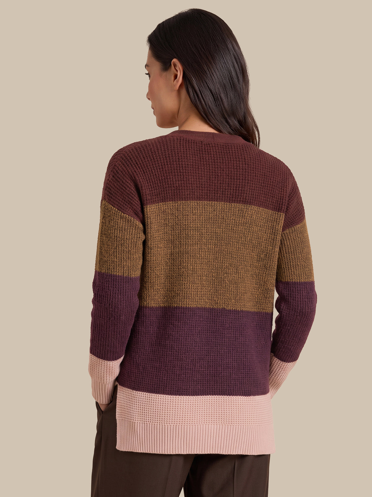 A woman shown from the back wearing a waffle-knit sweater with horizontal color-blocked stripes in dark burgundy, tan, and light pink, paired with dark brown pants against a solid beige background.
