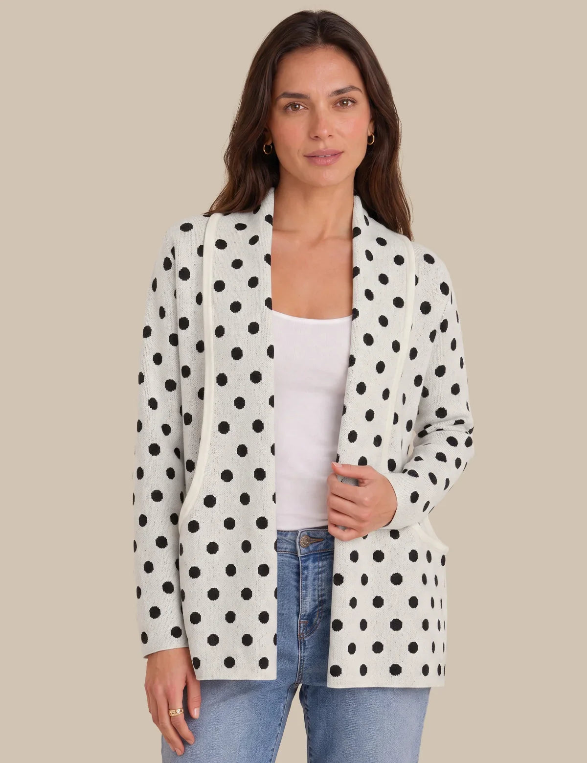 A woman modeling a white open-front blazer with black polka dots, worn over a white tank top and blue jeans, against a solid beige background.