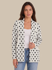A woman modeling a white open-front blazer with black polka dots, worn over a white tank top and blue jeans, against a solid beige background.