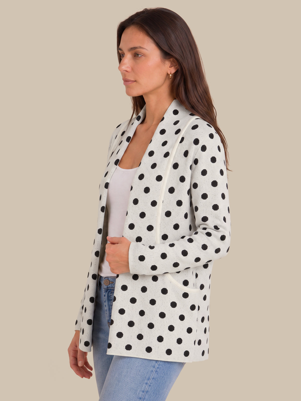 A woman wearing a white long-sleeve blazer with black polka dots, styled open over a white top and blue jeans, set against a solid beige background.