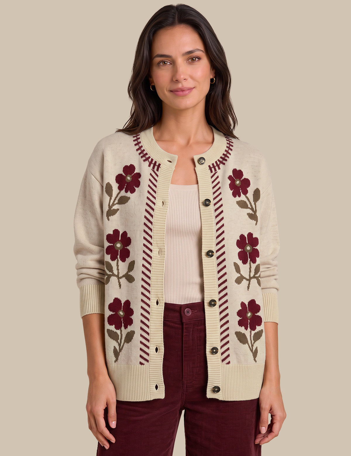 A woman modeling a cream-colored knit cardigan with burgundy floral and striped patterns, worn over a cream top and burgundy pants against a solid beige background.