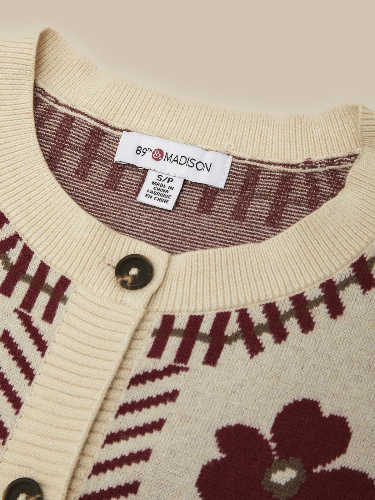 Close-up of a cream-colored knit cardigan sweater with burgundy and olive green patterns, featuring a visible '89° MADISON' brand label inside the collar.