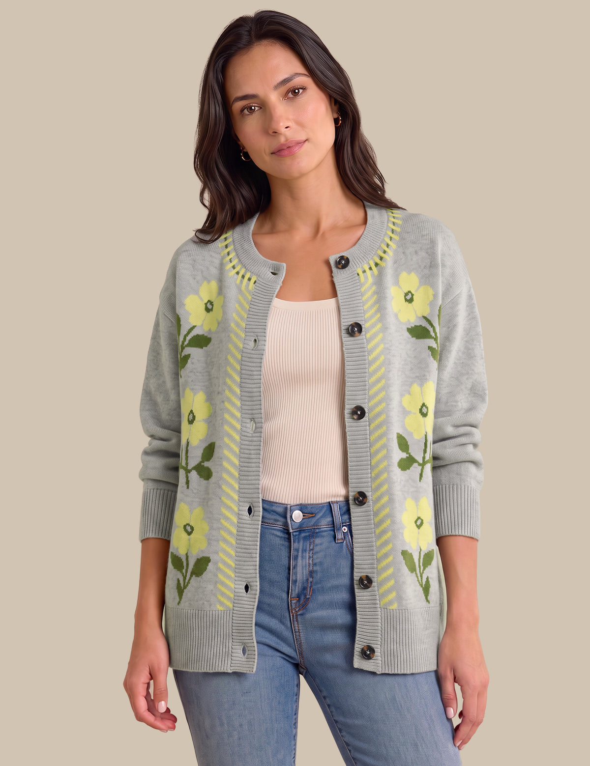 A woman modeling a light gray knit cardigan with yellow floral patterns and green leaves, worn over a cream ribbed top and blue jeans, against a solid beige background.