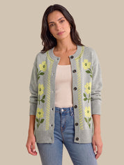 A woman modeling a light gray knit cardigan with yellow floral patterns and green leaves, worn over a cream ribbed top and blue jeans, against a solid beige background.